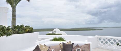 A white outdoor seating area with cushions and a tray of drinks, overlooking a body of water and palm trees.