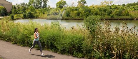 A woman jogging along a paved path beside a body of water with lush greenery and trees in the background.