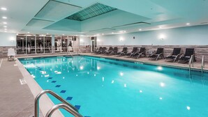 Une piscine intérieure avec des lignes de démarcation bien visibles, un vestiaire doté d’un mur entièrement recouvert de miroirs, et une rangée de chaises longues disposées le long du bord de la piscine.