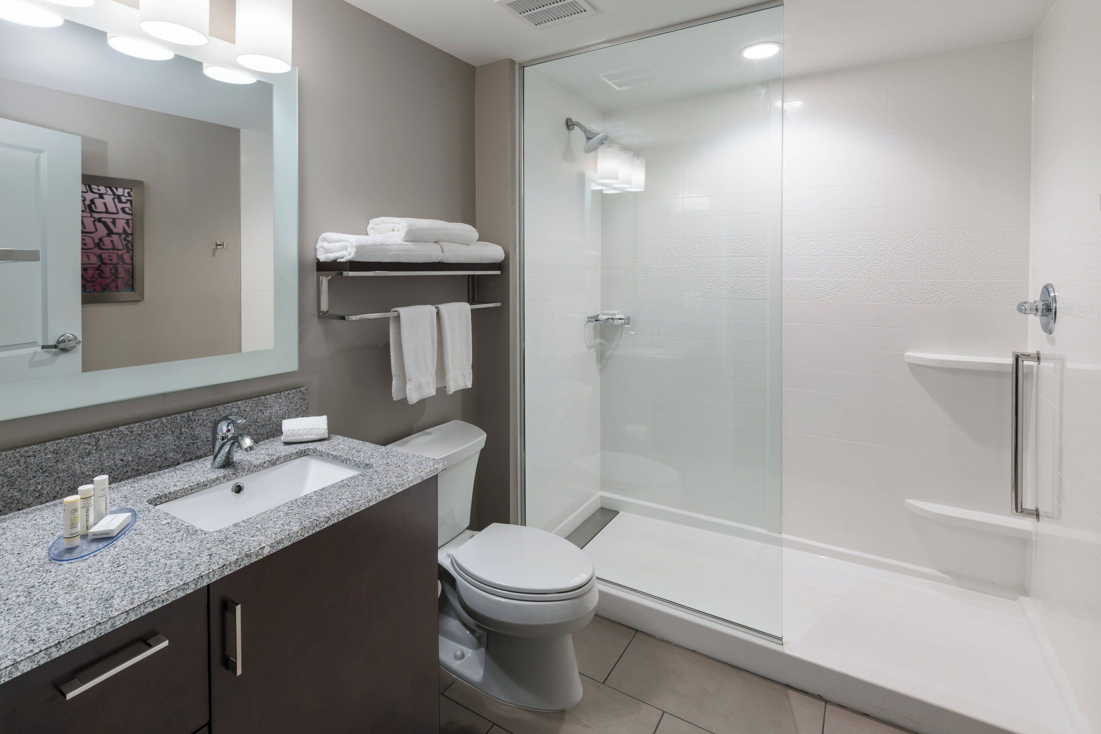 Un baño moderno con encimera de granito, inodoro, ducha con mampara de vidrio y espejo sobre el lavabo.