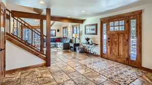 A spacious hallway with a wooden staircase, a living area with a sofa and a coffee table, and a large wooden door with glass panels.