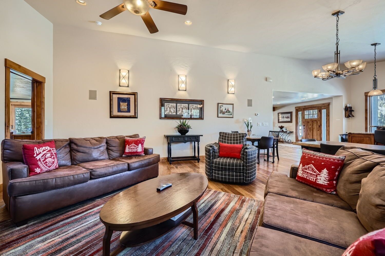 A spacious living room with a brown leather sofa, a plaid armchair, and a wooden coffee table. There are framed pictures on the walls and a ceiling fan.