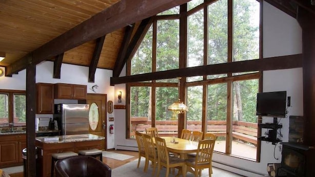 A spacious living area with a wooden ceiling, large windows, a dining table with chairs, a fireplace, and a kitchen with modern appliances.
