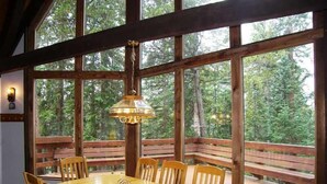 A wooden dining table with six chairs, a hanging light fixture, and large windows with a view of trees.