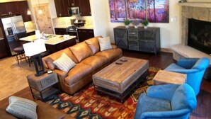 Ein modernes Wohnzimmer mit einem Ecksofa, einem Couchtisch und blauen Sessel. Im Hintergrund befindet sich eine Küche mit dunklen Schränken und einer Mikrowelle. Am rechten Rand ist ein Kamin zu sehen.