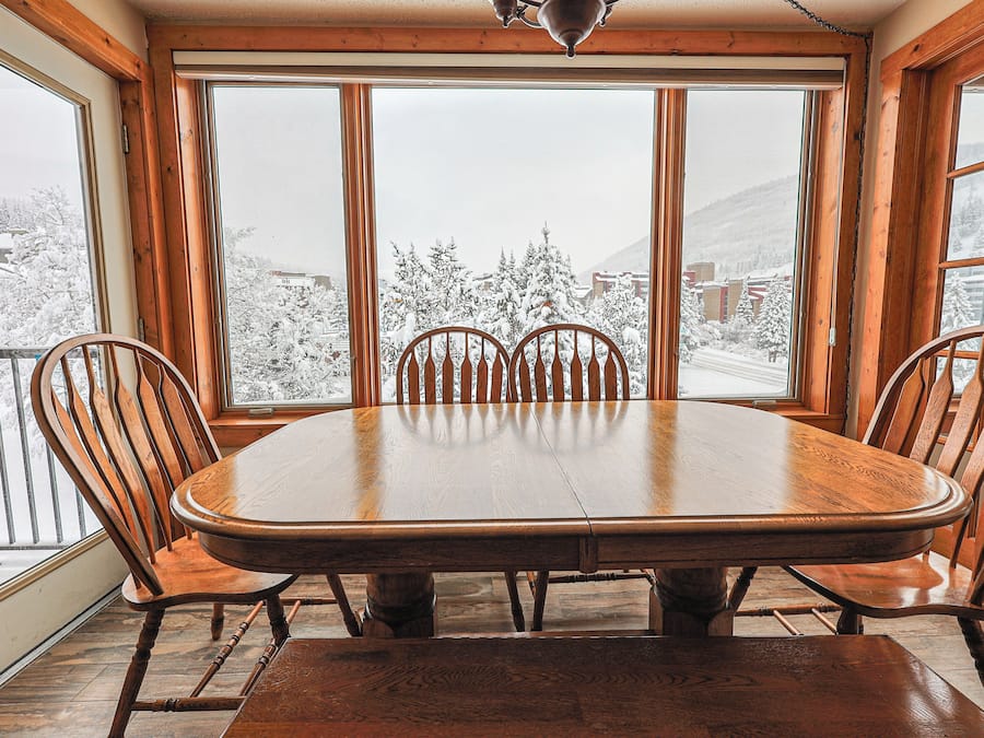 A dining area with a large wooden table and six chairs, large windows with a view of a snowy landscape.