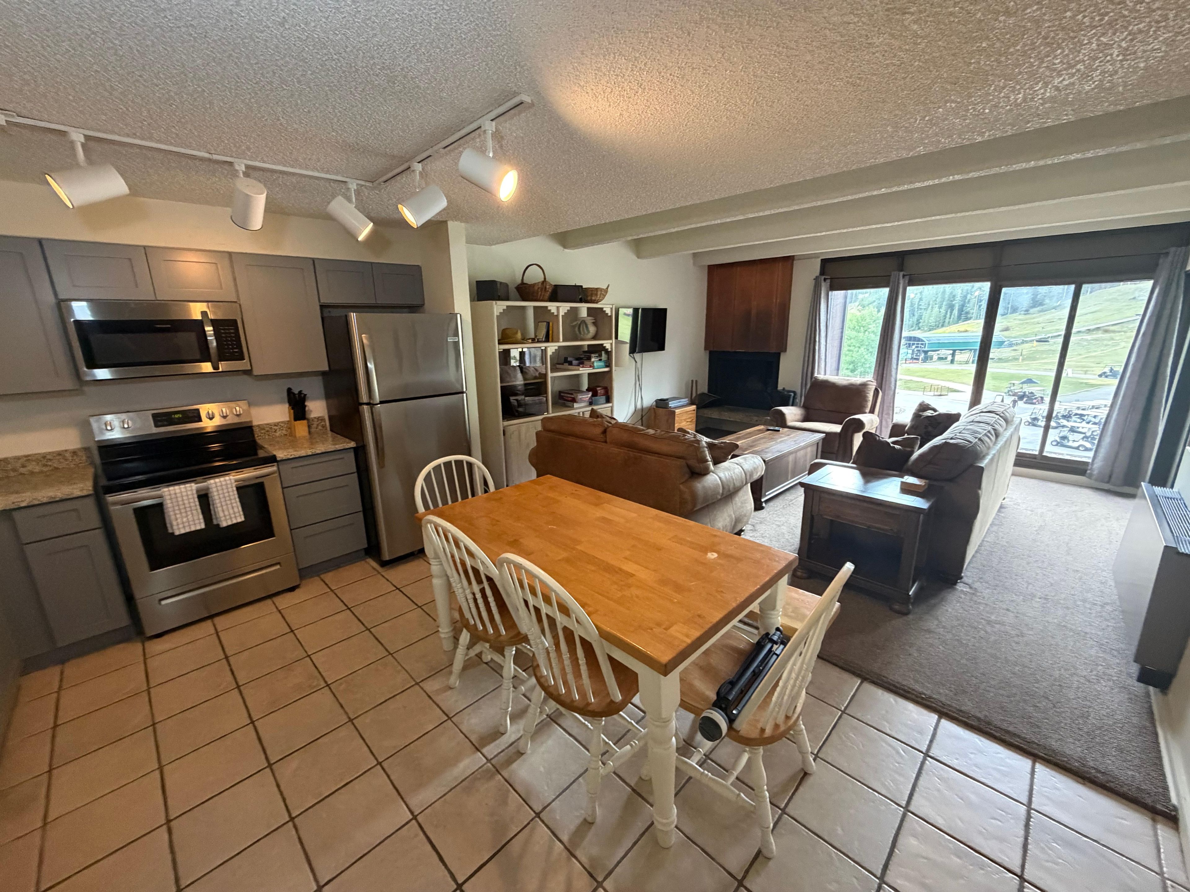 A modern kitchen with stainless steel appliances, a dining area with a wooden table and white chairs, and a living room with a brown sofa and a large window offering a view of the outdoors.