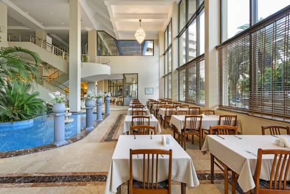 Un vestíbulo de hotel con piscina, varias zonas de comedor y una escalera que lleva a un nivel superior.