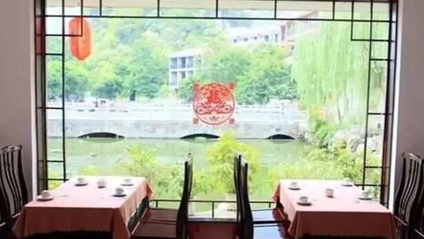 A dining area with tables set for two, a view of a river, and a decorative element hanging from the window. - Li River (Guilin)