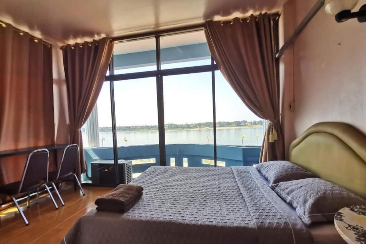 Quarto com uma cama grande, uma escrivaninha e uma cadeira, com vista para a água através de grandes janelas.