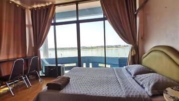 A bedroom with a large bed, a desk, and a chair, with a view of a body of water through large windows.