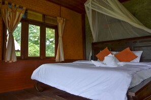 A bed with white linens and orange pillows, a wooden headboard, and a window with curtains.
