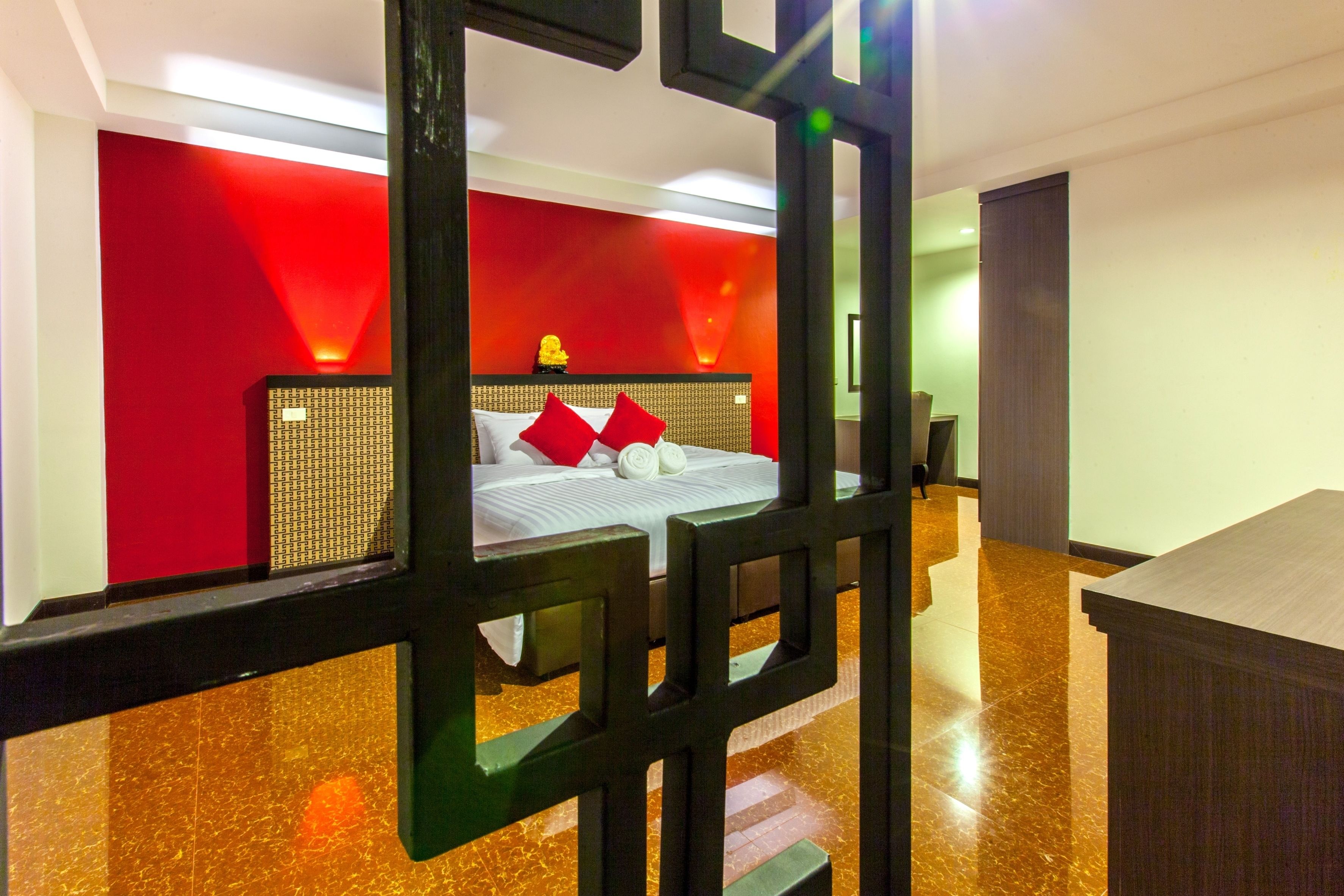 A hotel room with a red accent wall, a bed with white bedding and red pillows, a woven headboard, and a wooden desk.