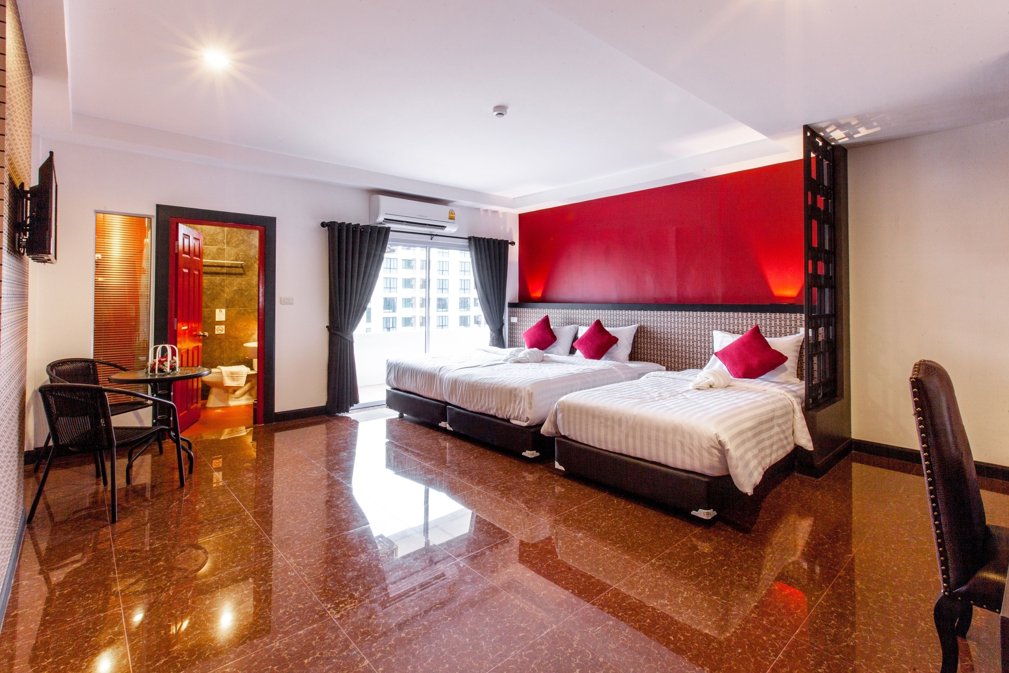 A hotel room with two beds, a sitting area with a chair and table, a red accent wall, and a view of the city through the window.