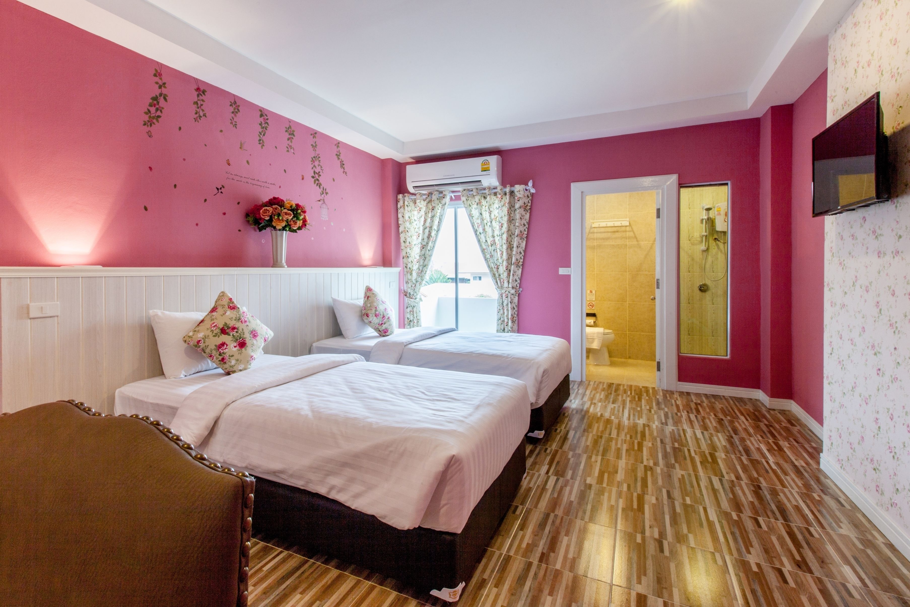 A hotel room with two beds, a wooden floor, a pink wall, and a TV mounted on the wall.