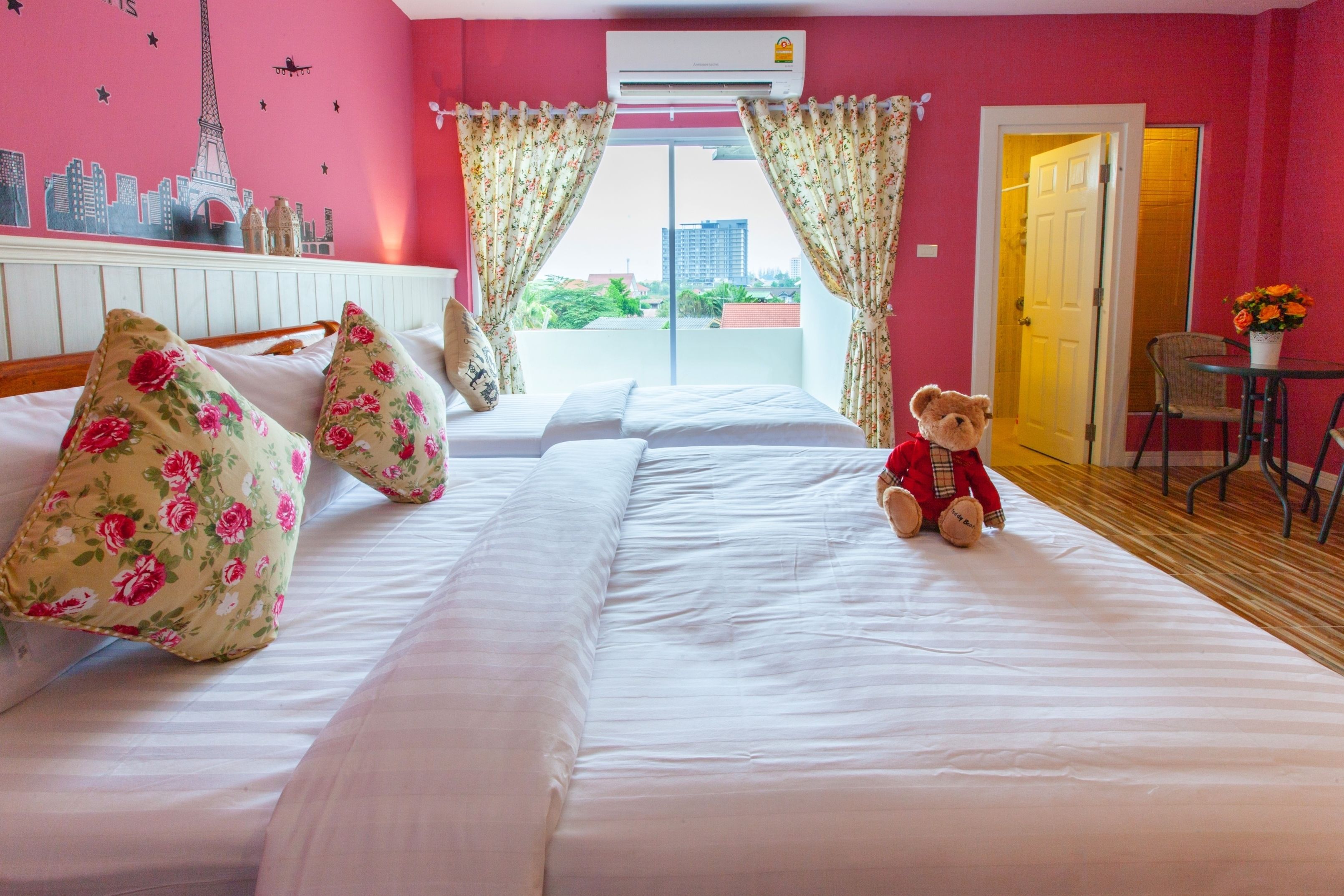 A hotel room with a bed, floral pillows, a teddy bear, a cityscape wall, and a view of buildings through the window.