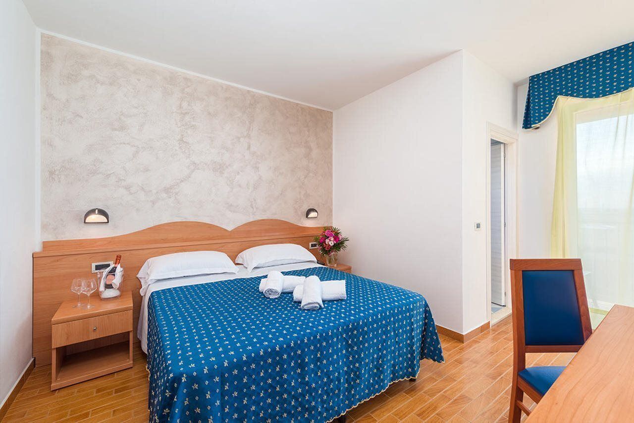 Una habitación de hotel con una cama de madera, una mesita de noche con un vaso y una botella, una colcha azul y una silla cerca de la ventana.