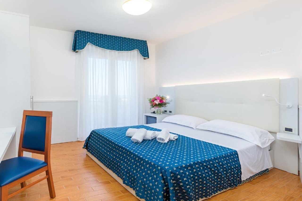 A hotel room with a bed, a chair, a nightstand with a vase of flowers, and a window with curtains.