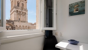 A room with a window overlooking a historic tower, a small table with a blue bowl, and a framed picture on the wall. - PLR Accommodation (Split)