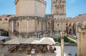 An outdoor café with tables and umbrellas, set against historic stone buildings with arches and columns. - PLR Accommodation (Split)