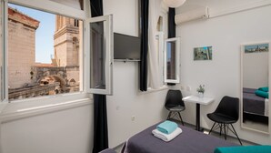 A hotel room with two beds, a small table, a chair, a TV, and a window with a view of a historic building. - PLR Accommodation (Split)