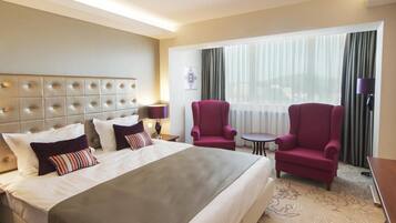 Ein Hotelzimmer mit einem großen Bett, zwei Sesseln, einem kleinen Tisch und einem Fenster mit Vorhängen.