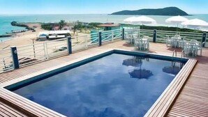 A rectangular swimming pool with a wooden deck surrounding it, overlooking a beach and ocean. - Hotel Bandeirantes (Balneario Barra do Sul)