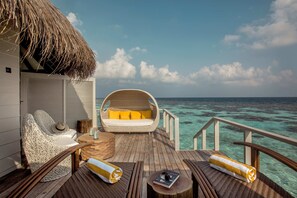 A wooden deck with a wicker chair, a cushioned seating area, and a thatched roof structure.