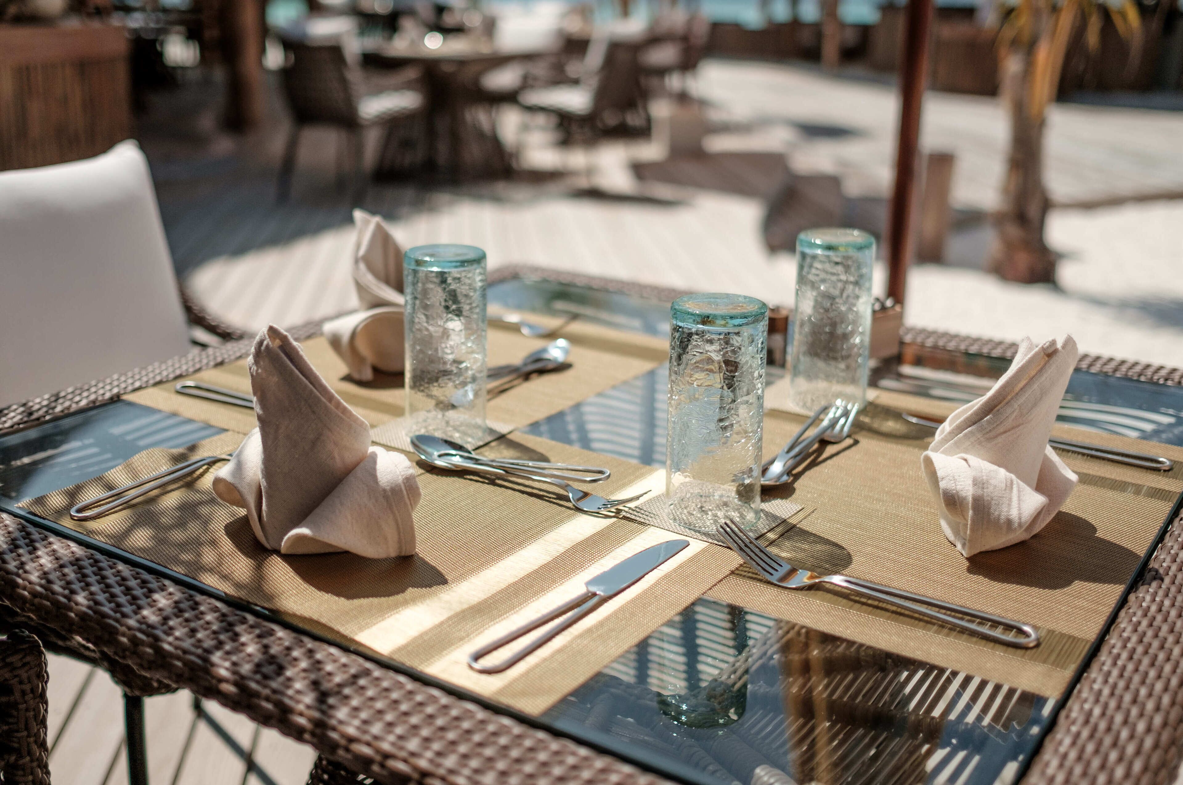 A dining table set with glassware, cutlery, and napkins on a glass table with a woven mat underneath.