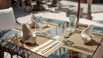 A dining table set with glassware, cutlery, and napkins on a glass table with a woven mat underneath.