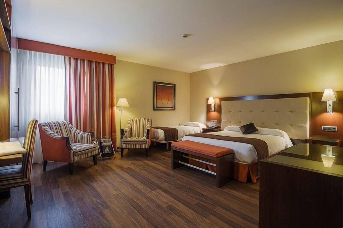 Une chambre d’hôtel avec deux lits, un bureau, une chaise, un canapé et une grande fenêtre avec des rideaux.