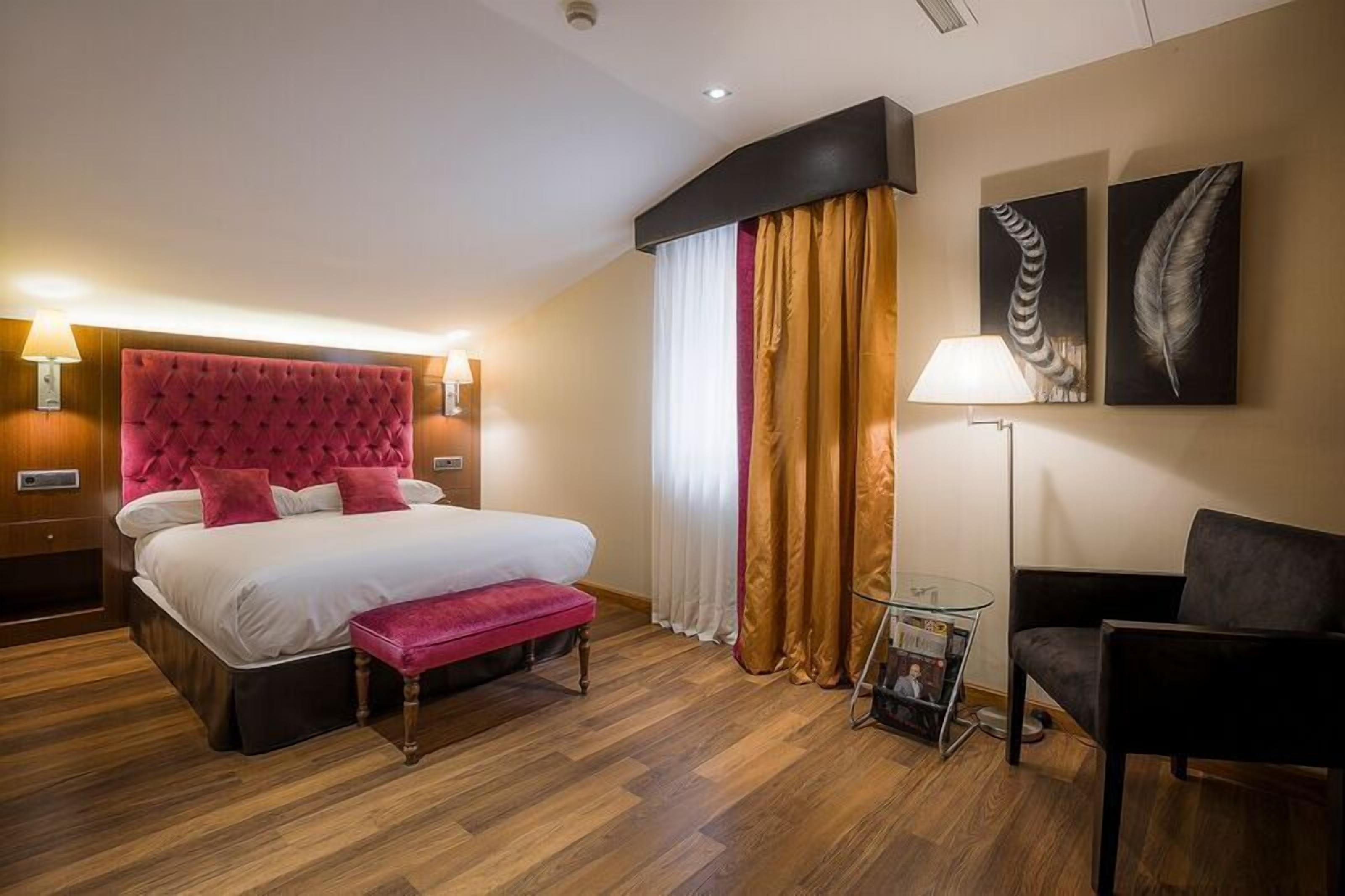 Habitación de hotel con una cama grande, un cabecero tapizado rojo, una mesita de noche con una lámpara y una silla.