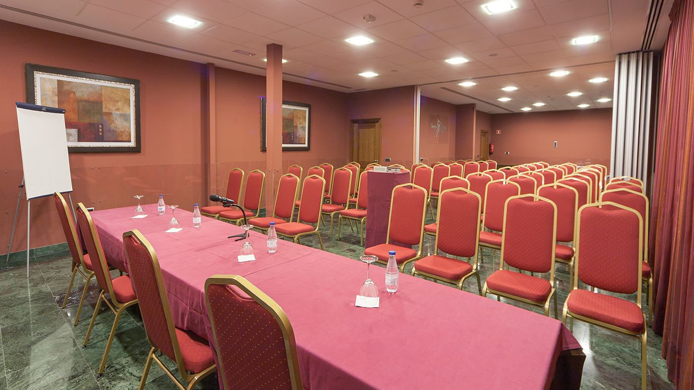 Un salón de conferencias con filas de sillas rojas, una mesa larga con mantel rojo y una pizarra.