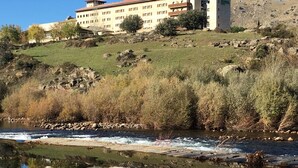 A building with multiple floors, a flat roof, and a reflection in a calm body of water. - Hospedium Hotel Mirador De Gredos (Barco De Avila)