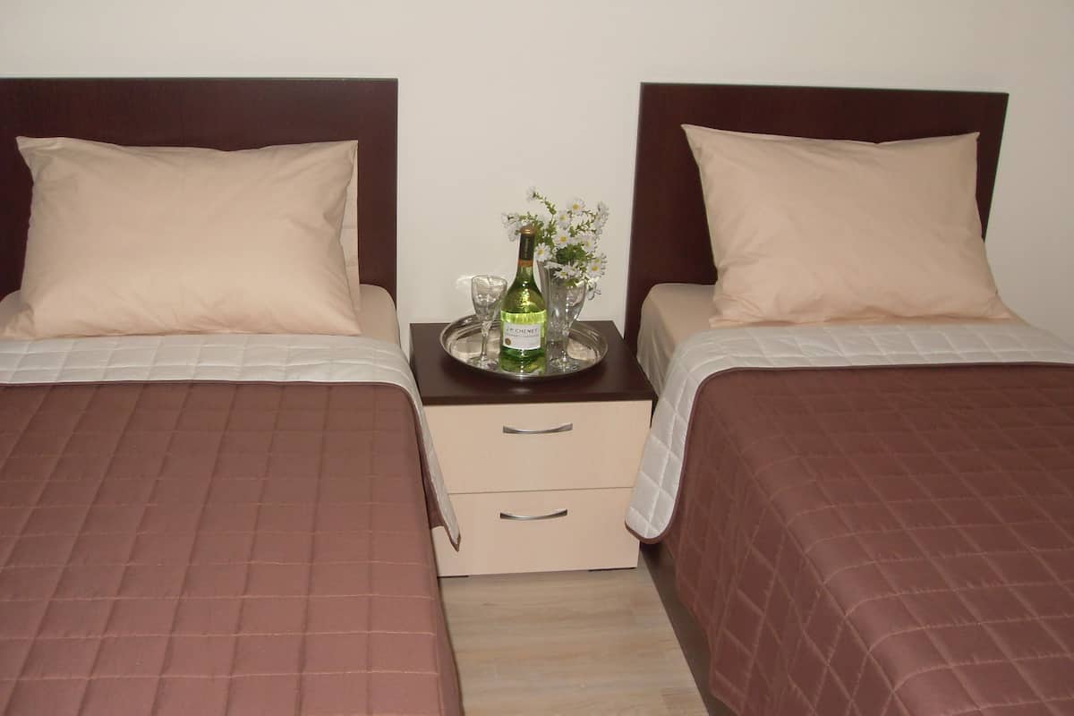 A hotel room with two beds, each with a brown quilted bedspread and a beige pillow. There is a nightstand between the beds with a tray holding a bottle of champagne, a glass, and a vase with flowers.