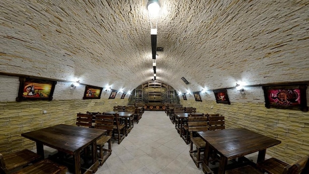 A long, narrow room with wooden tables and benches arranged in rows, a high ceiling with a central light fixture, and framed pictures on the walls. - Vila Lolo (Chisinau)