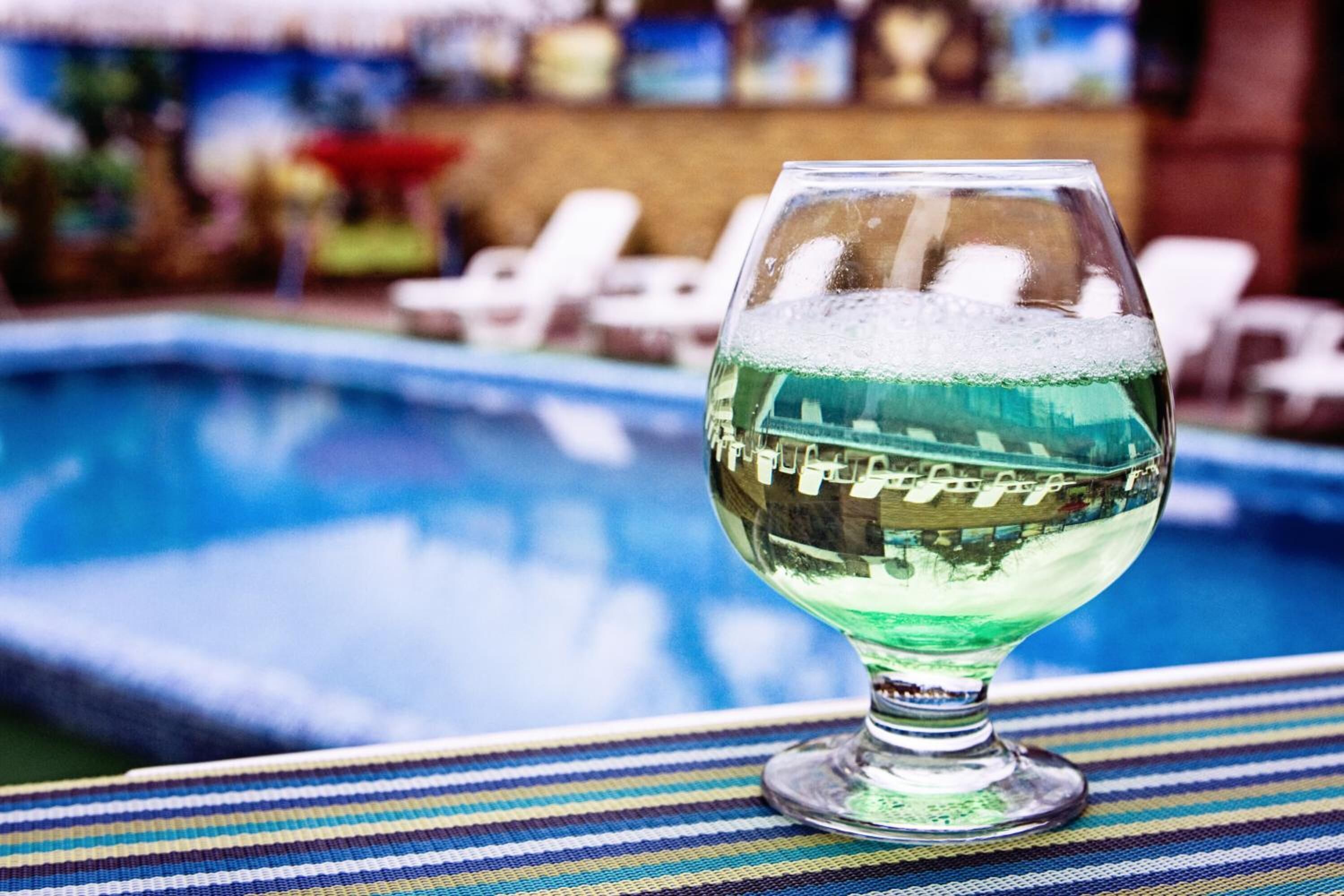 Ett glas med grön vätska står på ett randigt bord nära en pool.