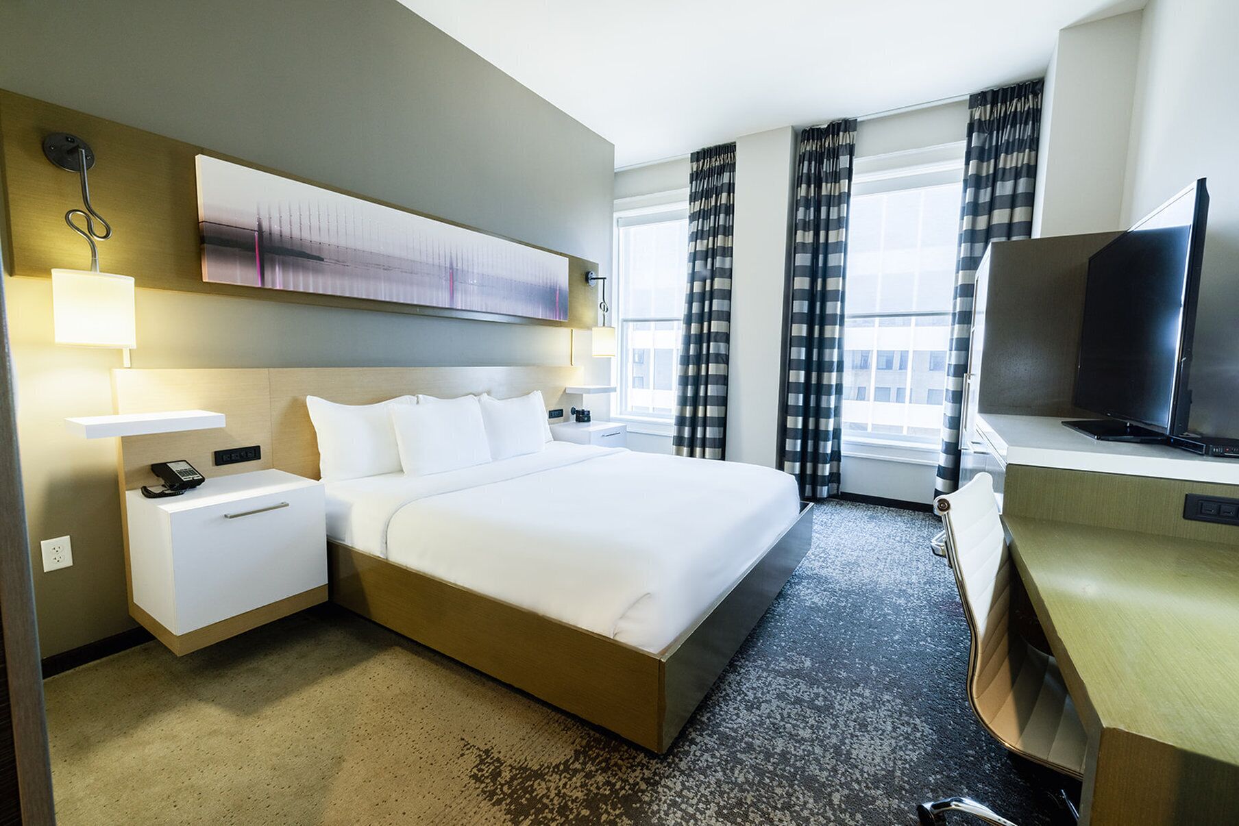 A hotel room with a bed, bedside table, lamp, TV, and a window with curtains.