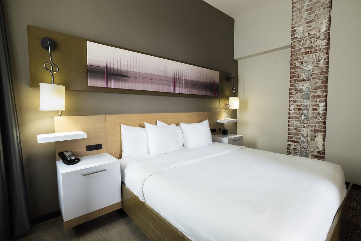 A hotel room with a large bed, a wooden headboard, a flat-screen TV mounted on the wall, a bedside table with a lamp, and a brick accent wall.