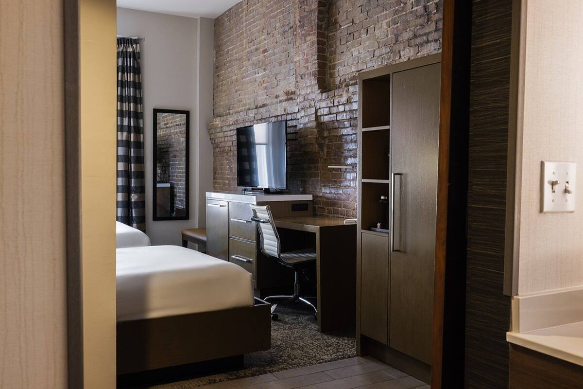 A compact hotel room with a brick accent wall, a bed, a desk with a chair, a TV, and a wardrobe.