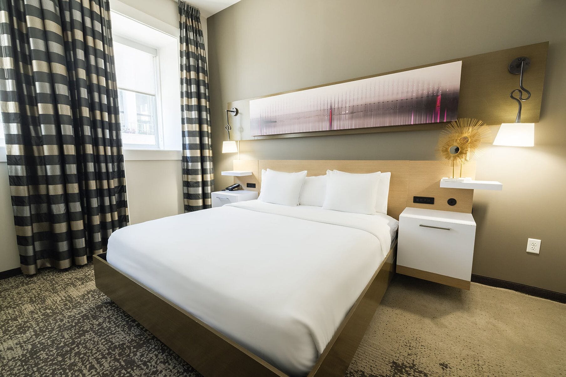 A hotel room with a large bed, a nightstand, a lamp, and a painting on the wall.