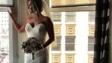 A bride in a wedding dress standing by a window with a view of a building.