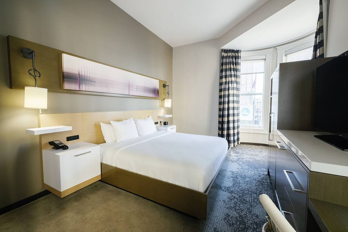 A hotel room with a large bed, bedside tables, a desk with a chair, a television, and a window with curtains.