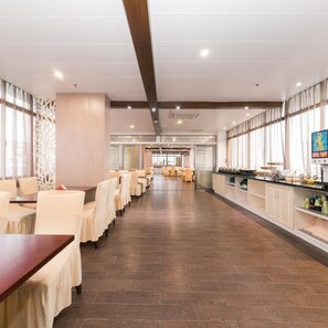 Ein Speisesaal eines Hotels mit Tischen und Stühlen, eine lange Theke mit einem Buffet und Blick durch Glastüren.
