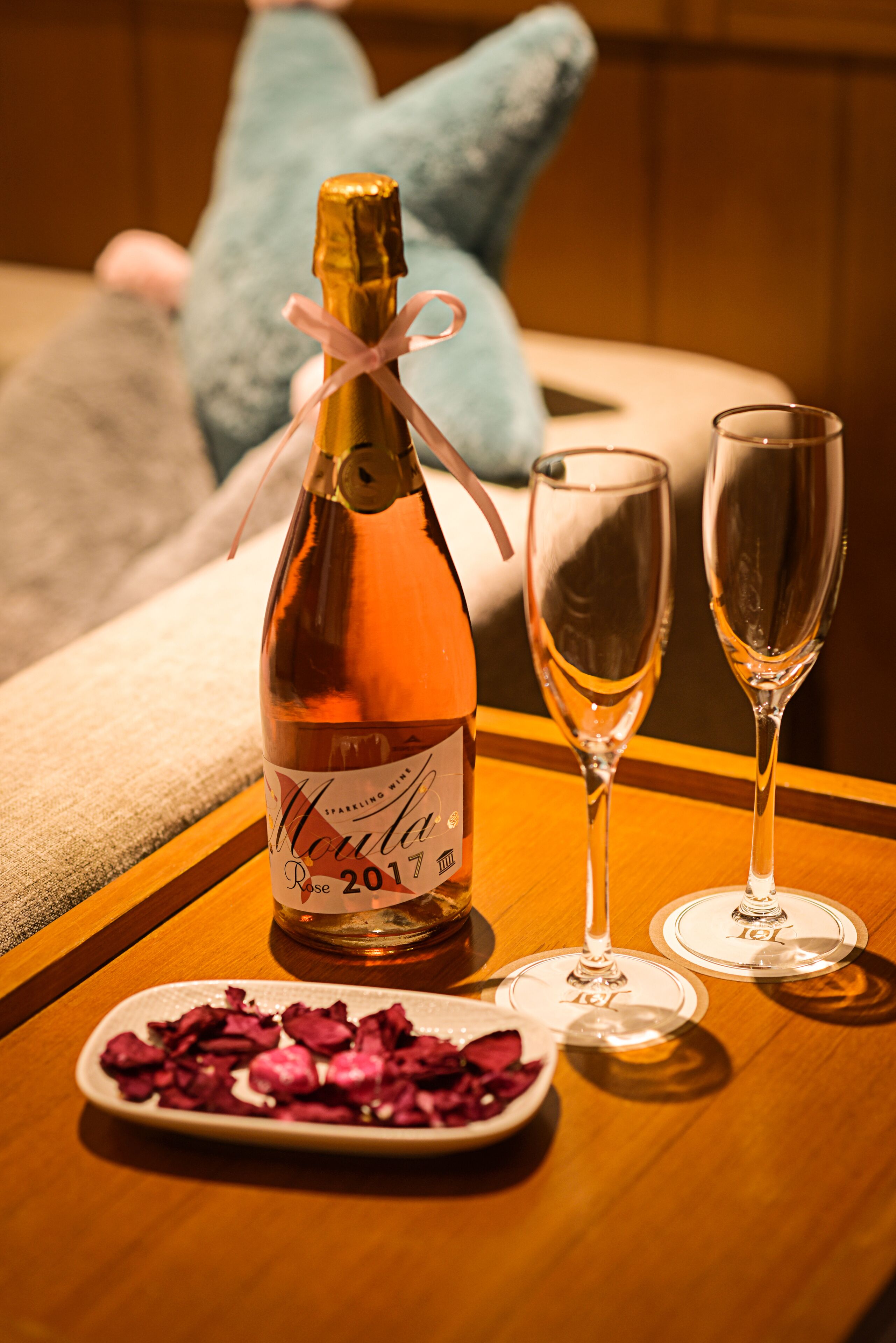 A bottle of wine with a label, two empty wine glasses, and a plate of rose petals on a wooden table.