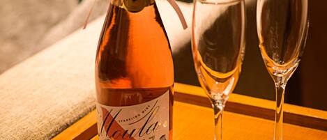 A bottle of wine with a label, two empty wine glasses, and a plate of rose petals on a wooden table.