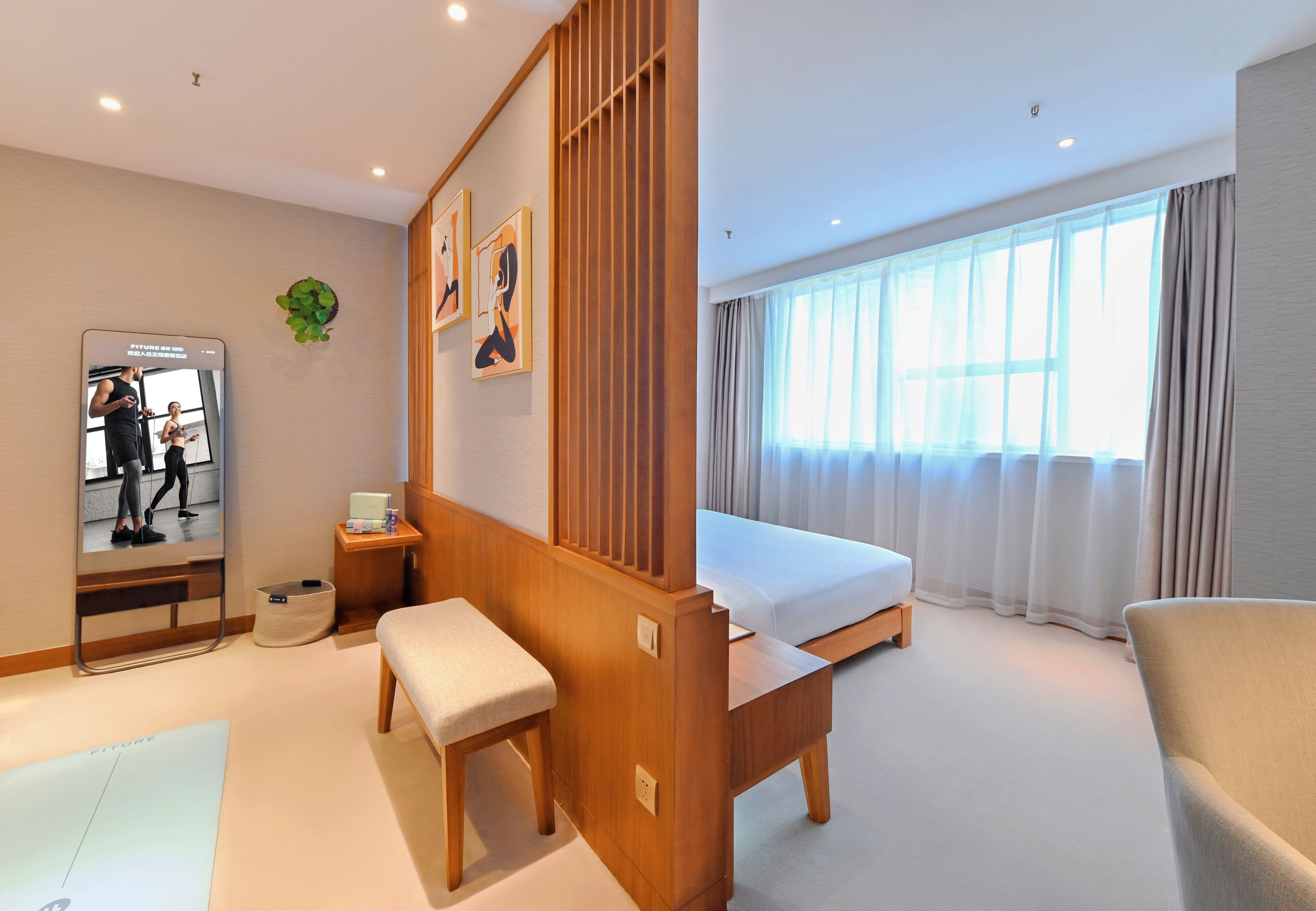 A hotel room with a bed, a wooden desk, a bench, a mirror, and a large window with curtains.