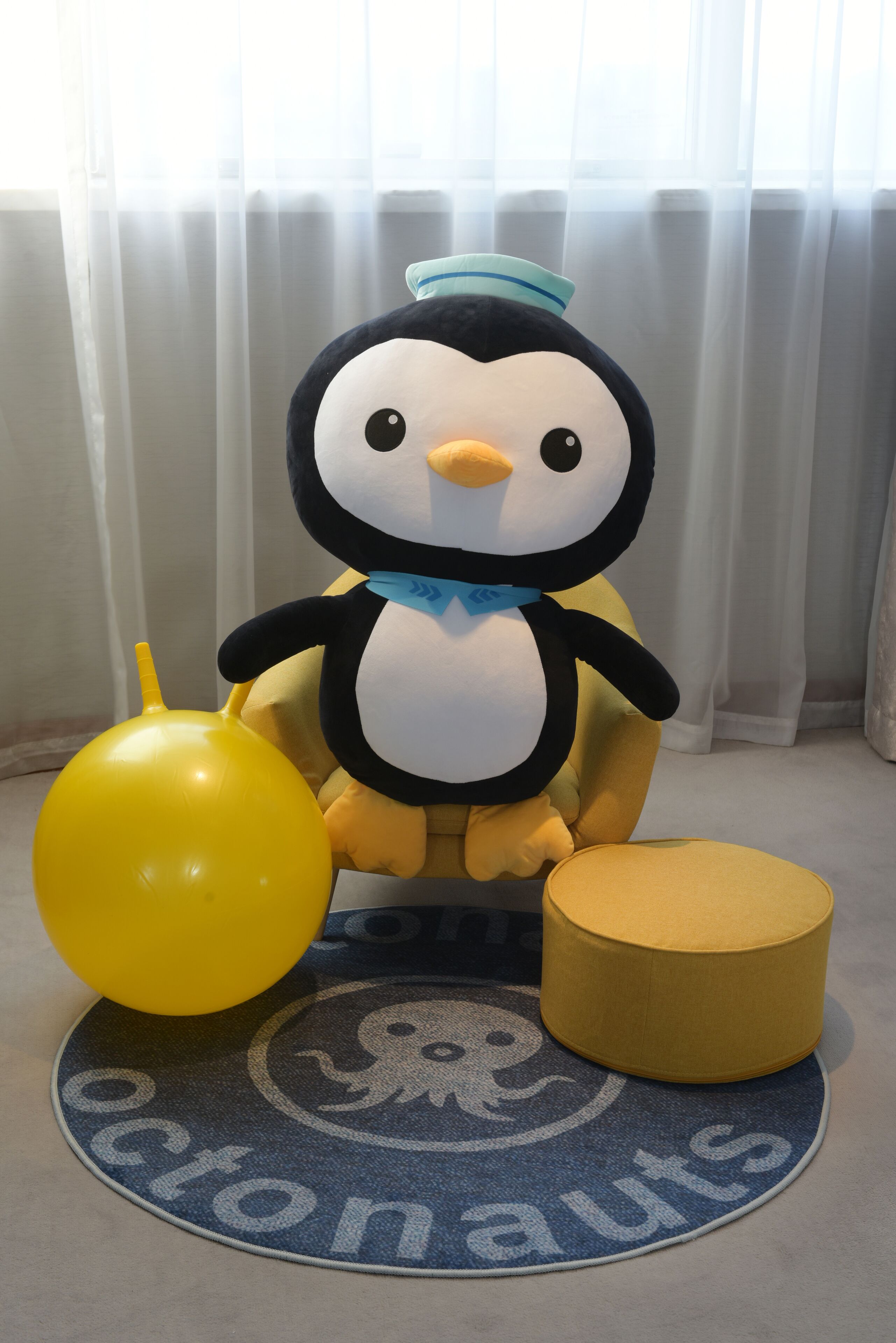 Un jouet en peluche représentant un pingouin, avec un ballon jaune et un coussin rond, posé sur un tapis rond orné d’un motif circulaire.
