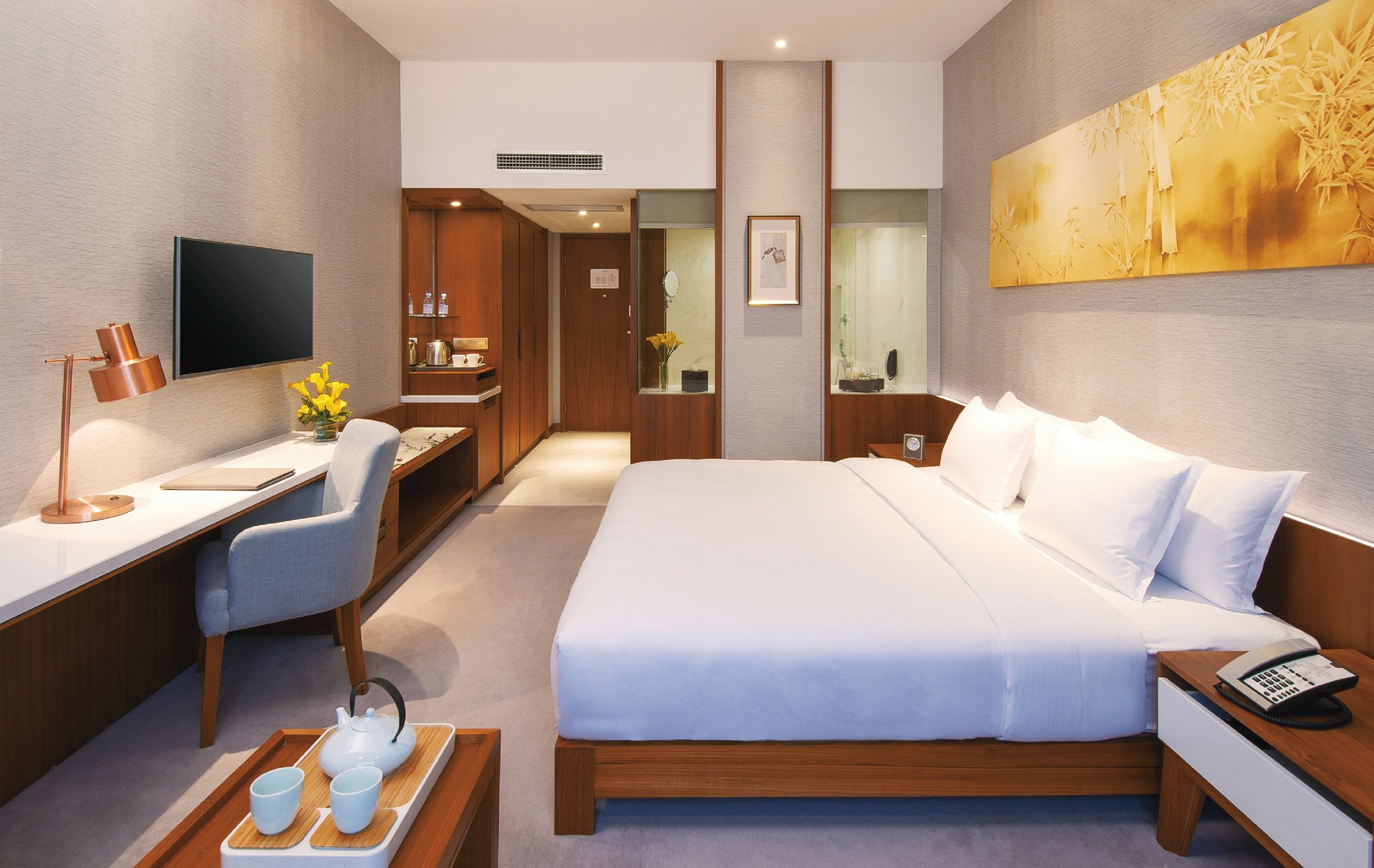 A modern hotel room with a large bed, a desk with a chair, a television, and a bathroom visible through a glass door.