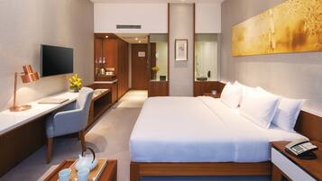 A modern hotel room with a large bed, a desk with a chair, a television, and a bathroom visible through a glass door.